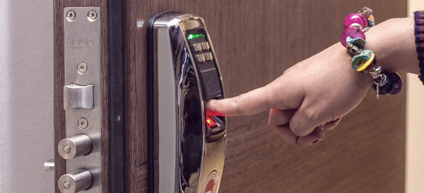 A finger held against the lit fingerprint reader of a smart lock on a wooden door, with the door's steel multipoint bolts visible along its open edge.