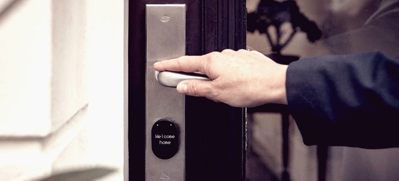 A hand on the lever of a stainless smart lock fitted to a dark panelled front door, its small display lit.