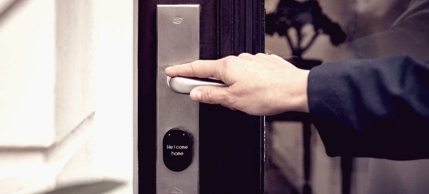 A hand on the lever of a stainless smart lock fitted to a dark panelled front door, its small display reading welcome home.